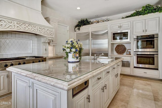 kitchen featuring custom exhaust hood, backsplash, built in appliances, an island with sink, and light stone counters