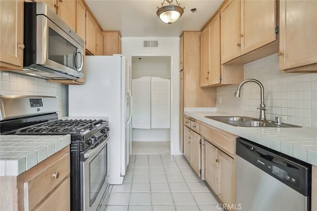 kitchen with tasteful backsplash, tile countertops, light brown cabinetry, and appliances with stainless steel finishes