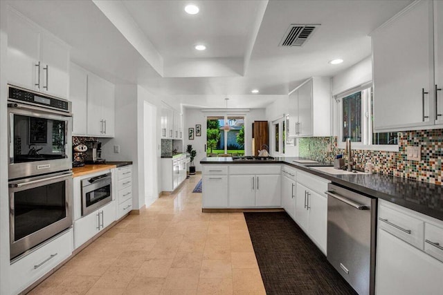 kitchen with kitchen peninsula, a raised ceiling, appliances with stainless steel finishes, backsplash, and white cabinetry