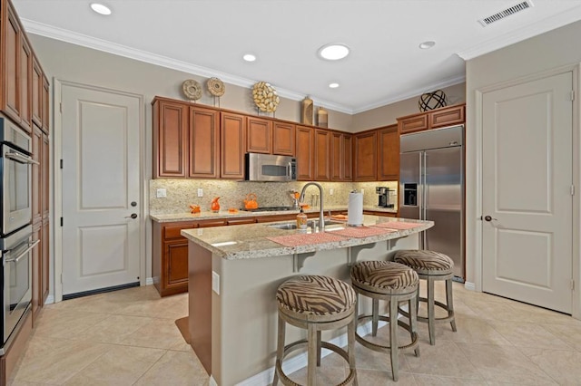 kitchen with sink, light stone counters, an island with sink, appliances with stainless steel finishes, and ornamental molding