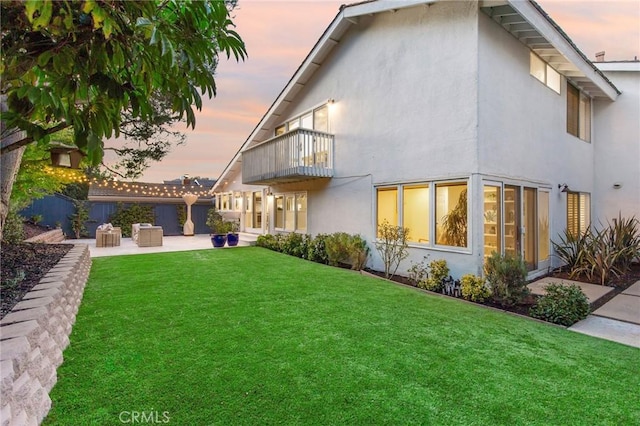 back house at dusk featuring a balcony, a patio area, and a lawn