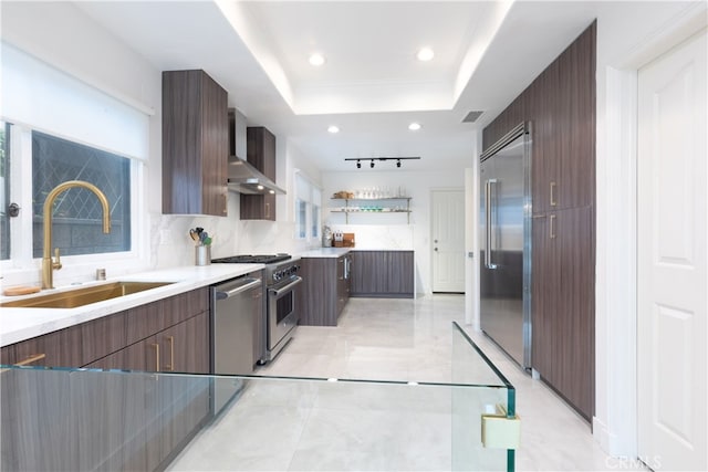 kitchen featuring a raised ceiling, backsplash, stainless steel appliances, wall chimney exhaust hood, and sink