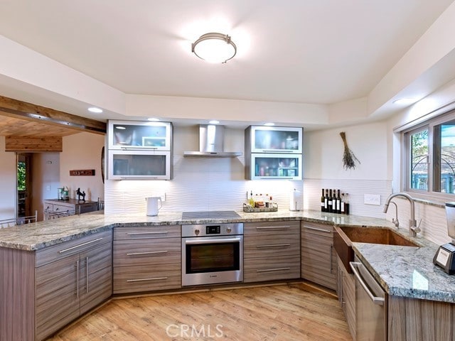 kitchen with wall chimney range hood, light stone countertops, light hardwood / wood-style flooring, sink, and stainless steel appliances