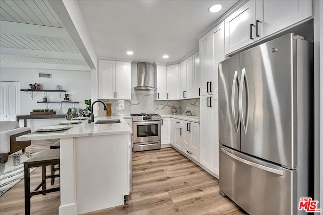 kitchen featuring wall chimney range hood, a kitchen breakfast bar, light hardwood / wood-style floors, white cabinets, and appliances with stainless steel finishes