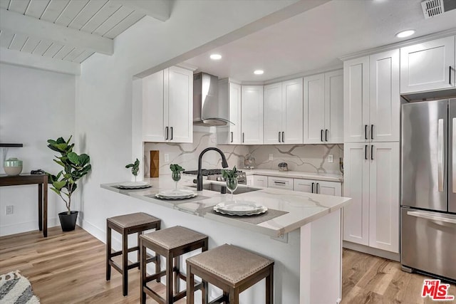 kitchen featuring wall chimney exhaust hood, appliances with stainless steel finishes, beamed ceiling, white cabinetry, and a breakfast bar area