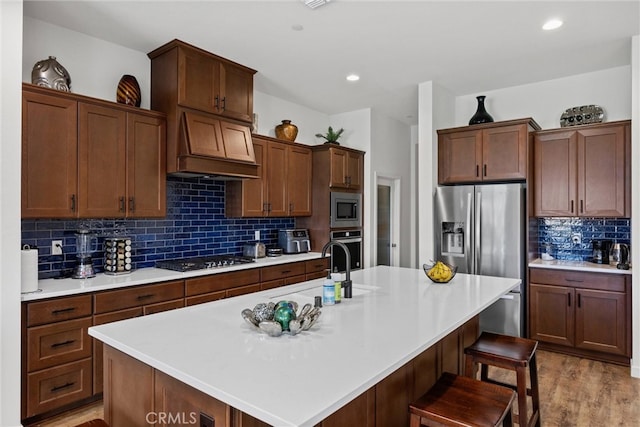 kitchen featuring custom range hood, appliances with stainless steel finishes, light wood-type flooring, and a kitchen island with sink