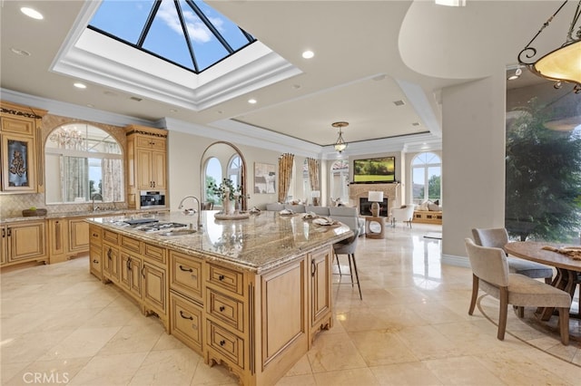 kitchen featuring decorative backsplash, light stone counters, a tray ceiling, crown molding, and an island with sink