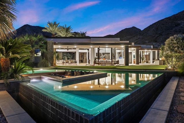 pool at dusk with outdoor lounge area, ceiling fan, a patio area, a mountain view, and a hot tub