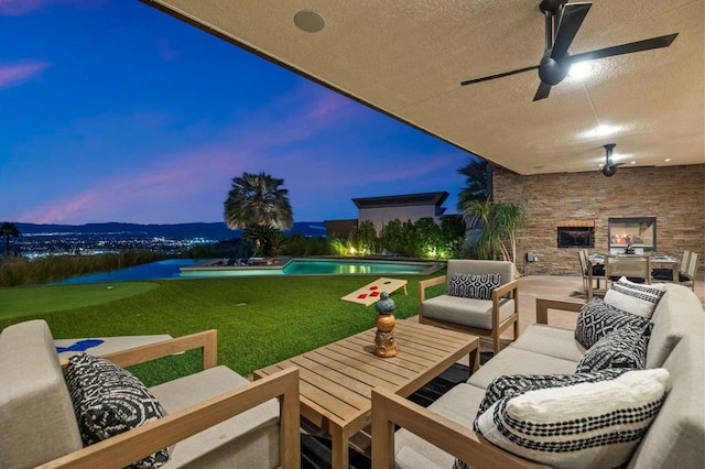 patio terrace at dusk with an outdoor living space with a fireplace, ceiling fan, and a lawn