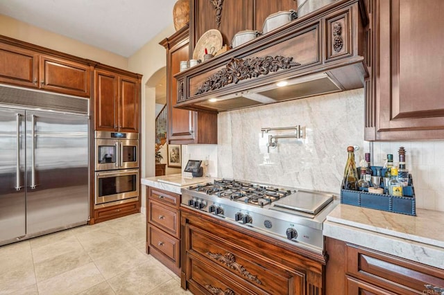 kitchen featuring decorative backsplash, appliances with stainless steel finishes, light stone countertops, custom range hood, and light tile patterned floors