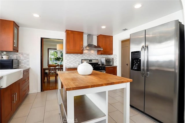 kitchen featuring appliances with stainless steel finishes, tasteful backsplash, wall chimney range hood, light tile patterned floors, and butcher block counters