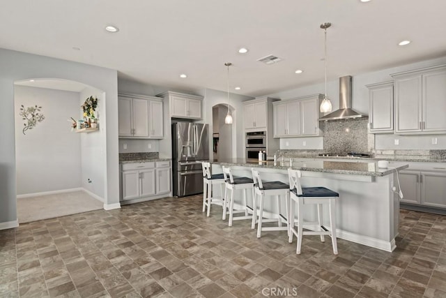 kitchen featuring wall chimney exhaust hood, light stone counters, a center island with sink, pendant lighting, and stainless steel appliances