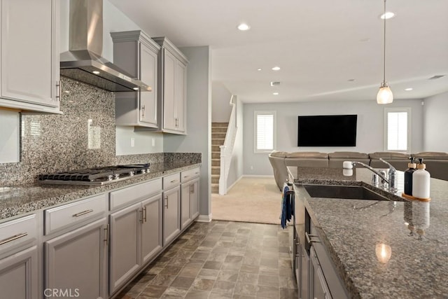 kitchen with sink, gray cabinetry, hanging light fixtures, dark stone counters, and wall chimney range hood