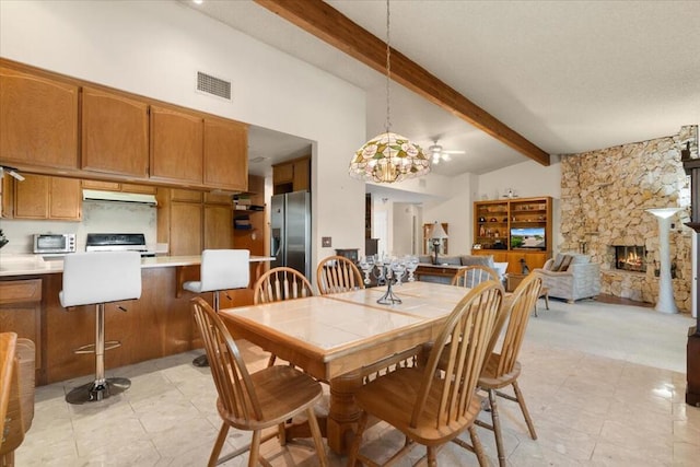 dining area featuring ceiling fan with notable chandelier, beam ceiling, a stone fireplace, and high vaulted ceiling