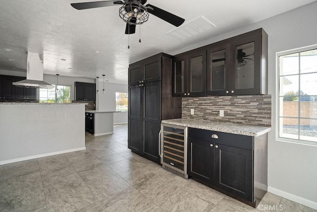 kitchen featuring light stone countertops, hanging light fixtures, wine cooler, decorative backsplash, and exhaust hood