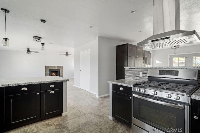 kitchen featuring stainless steel range with gas cooktop, hanging light fixtures, ceiling fan, range hood, and a tiled fireplace