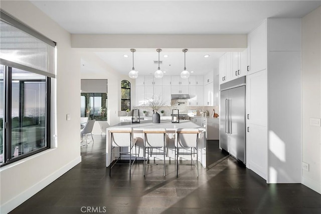 kitchen with under cabinet range hood, white cabinets, backsplash, stainless steel built in fridge, and a center island with sink