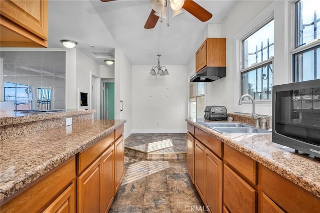 kitchen with light stone counters, brown cabinets, decorative light fixtures, a sink, and ceiling fan with notable chandelier