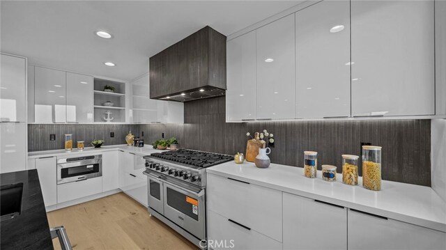 kitchen with backsplash, stainless steel appliances, wall chimney range hood, light hardwood / wood-style floors, and white cabinetry