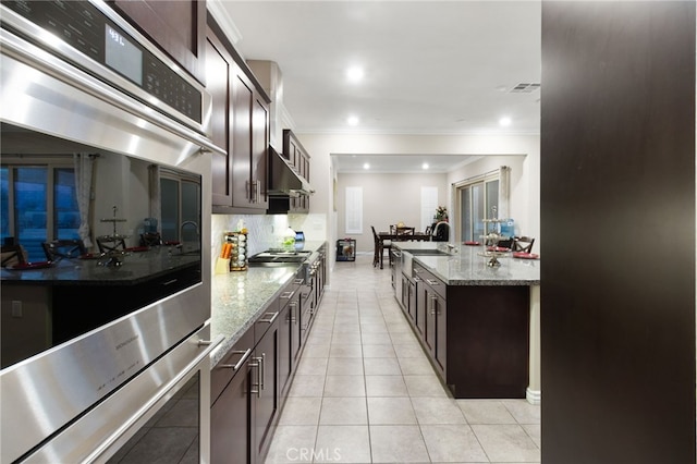 kitchen with dark brown cabinetry, light stone counters, light tile patterned floors, and ornamental molding