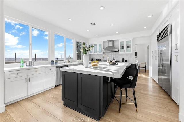 kitchen with stainless steel appliances, wall chimney range hood, white cabinets, light hardwood / wood-style floors, and an island with sink