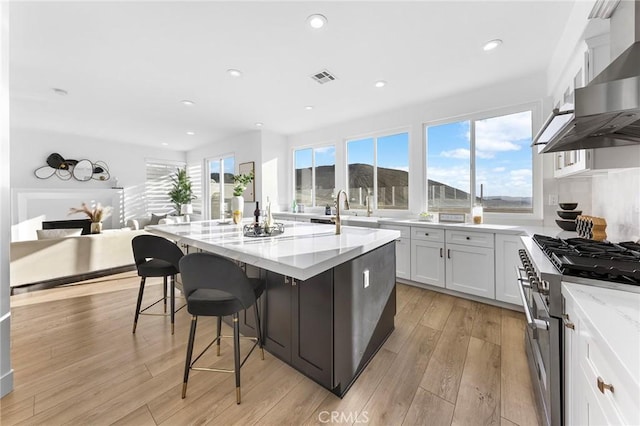 kitchen with a kitchen island with sink, white cabinets, high end stainless steel range oven, wall chimney exhaust hood, and light stone countertops