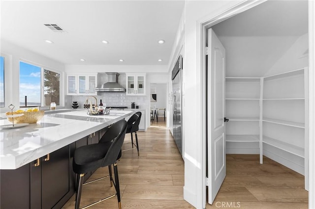 kitchen with light stone countertops, wall chimney range hood, tasteful backsplash, light hardwood / wood-style flooring, and white cabinets