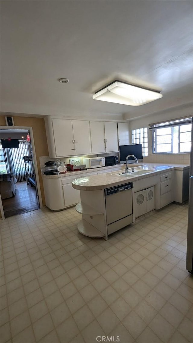 kitchen featuring sink, light tile patterned flooring, kitchen peninsula, dishwashing machine, and white cabinets