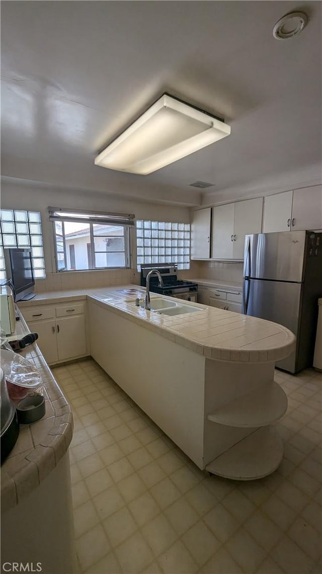 kitchen with sink, kitchen peninsula, tile counters, white cabinetry, and stainless steel refrigerator