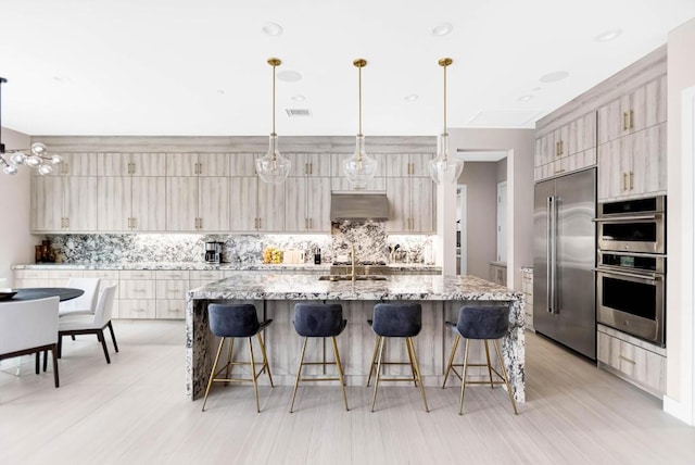 kitchen featuring appliances with stainless steel finishes, decorative light fixtures, light stone counters, exhaust hood, and a kitchen island with sink