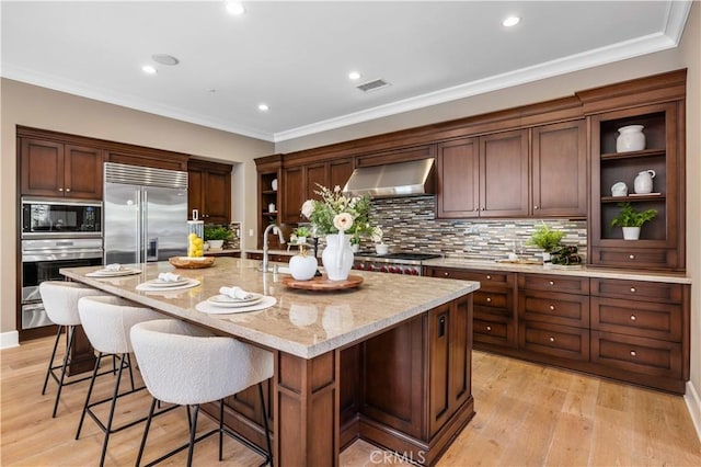 kitchen featuring light stone counters, wall chimney exhaust hood, built in appliances, light hardwood / wood-style floors, and an island with sink