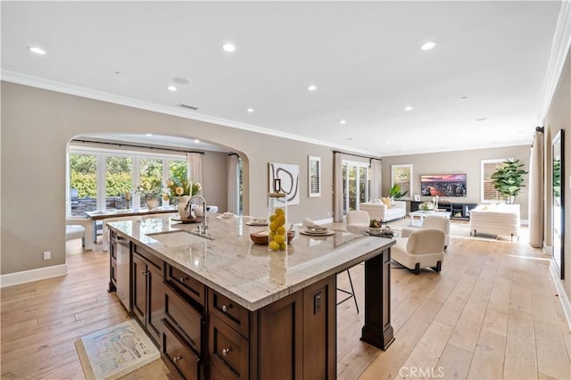 kitchen with light wood-type flooring, light stone counters, ornamental molding, sink, and a center island with sink