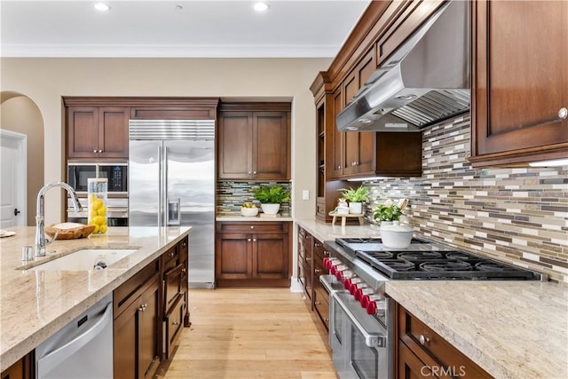 kitchen with light stone countertops, sink, wall chimney exhaust hood, light hardwood / wood-style flooring, and built in appliances