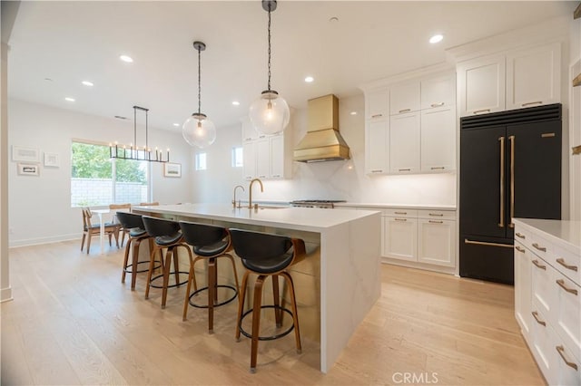 kitchen featuring built in fridge, decorative light fixtures, white cabinetry, custom exhaust hood, and a kitchen island with sink