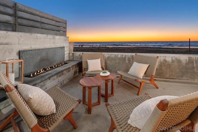 patio terrace at dusk featuring an outdoor stone fireplace, a water view, and a beach view