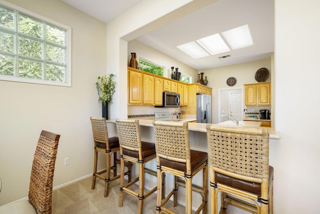 kitchen with light carpet, a skylight, appliances with stainless steel finishes, a kitchen bar, and kitchen peninsula