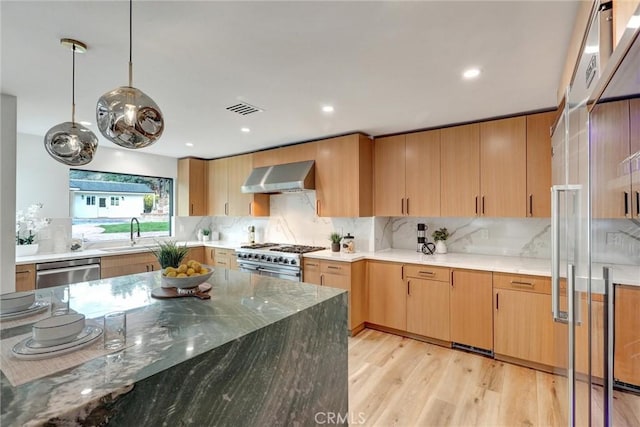 kitchen featuring dark stone counters, wall chimney exhaust hood, stainless steel appliances, decorative light fixtures, and light hardwood / wood-style floors