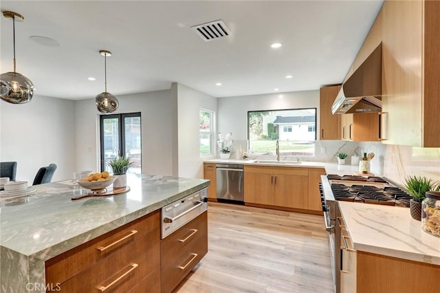 kitchen with wall chimney range hood, light stone counters, a spacious island, decorative light fixtures, and appliances with stainless steel finishes