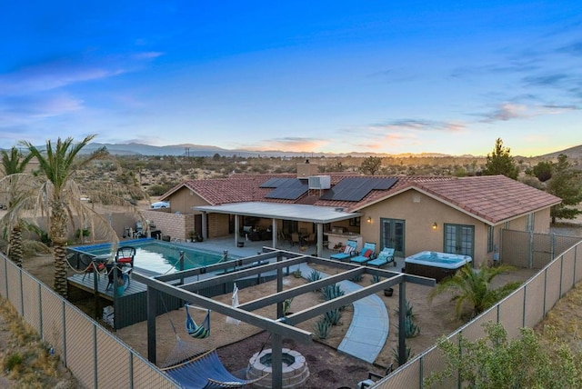 back house at dusk featuring a swimming pool with hot tub, solar panels, a patio, a mountain view, and an outdoor hangout area