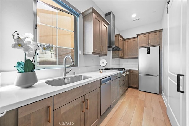 kitchen with wall chimney range hood, sink, appliances with stainless steel finishes, and light hardwood / wood-style flooring