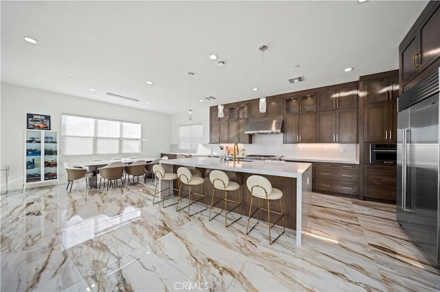 kitchen featuring built in fridge, dark brown cabinets, hanging light fixtures, and a kitchen island with sink