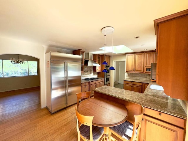 kitchen featuring appliances with stainless steel finishes, wall chimney exhaust hood, an inviting chandelier, backsplash, and light hardwood / wood-style flooring