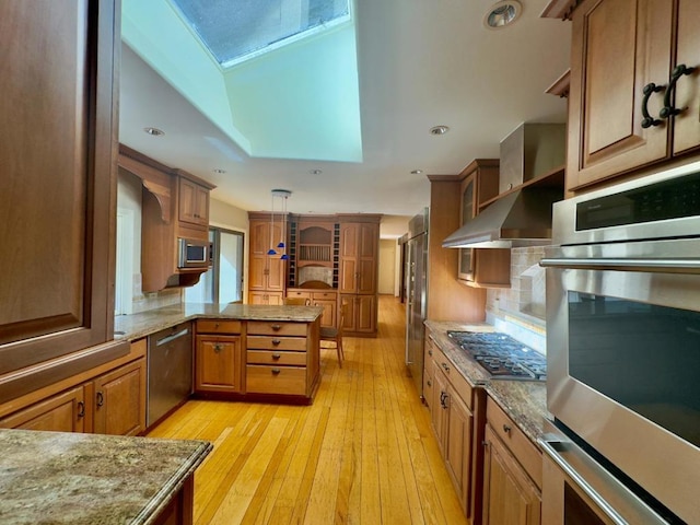 kitchen featuring a skylight, stainless steel appliances, decorative light fixtures, wall chimney range hood, and light hardwood / wood-style flooring