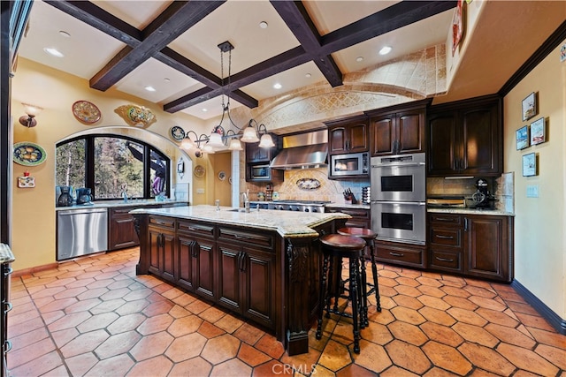 kitchen featuring wall chimney range hood, dark brown cabinetry, hanging light fixtures, a kitchen island with sink, and appliances with stainless steel finishes