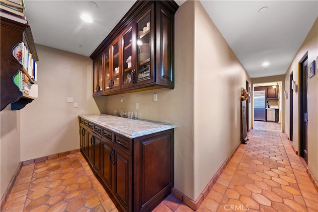 bar with light tile patterned floors, stainless steel built in fridge, light stone countertops, and dark brown cabinetry