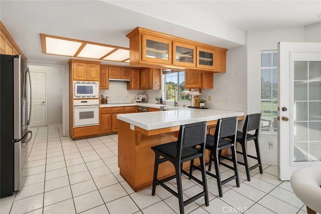 kitchen featuring kitchen peninsula, decorative backsplash, white appliances, light tile patterned floors, and a breakfast bar area