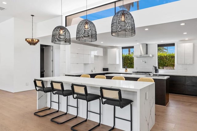 kitchen with light wood-type flooring, wall chimney exhaust hood, a spacious island, decorative light fixtures, and white cabinets