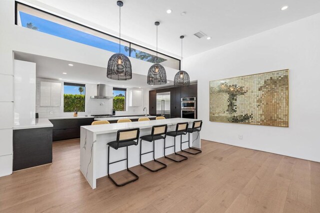 kitchen featuring a center island, wall chimney range hood, hanging light fixtures, light wood-type flooring, and stainless steel appliances