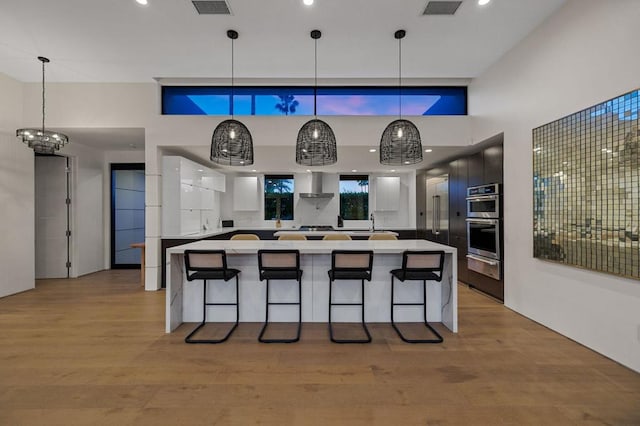 kitchen with hanging light fixtures, wall chimney exhaust hood, double oven, light hardwood / wood-style floors, and a breakfast bar area