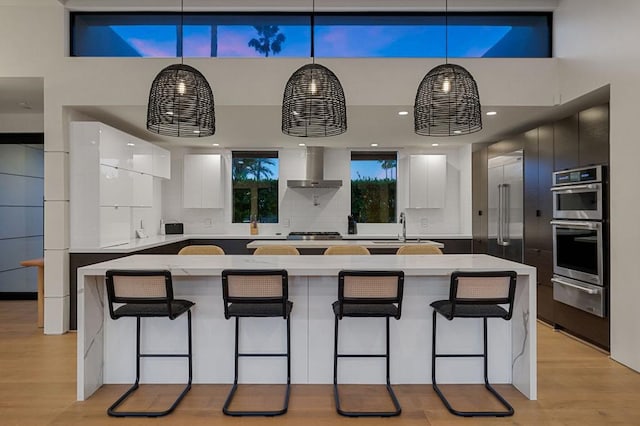 kitchen with white cabinetry, a large island, wall chimney range hood, light hardwood / wood-style floors, and decorative light fixtures